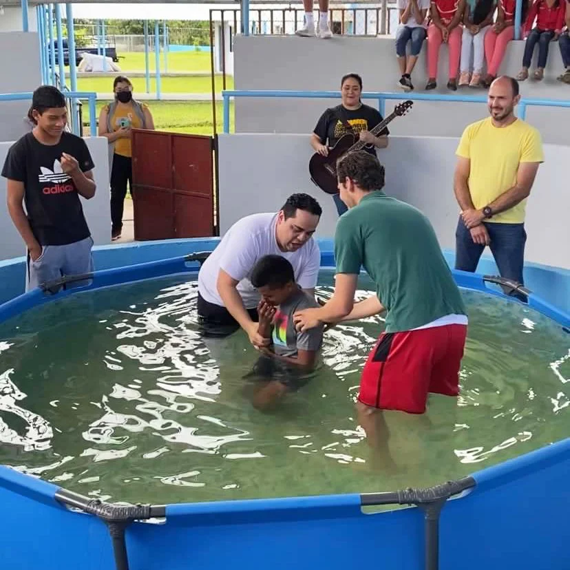 A person baptizing a young boy in a swimming pool during a religious ceremony, with onlookers and a woman playing guitar in the background.
