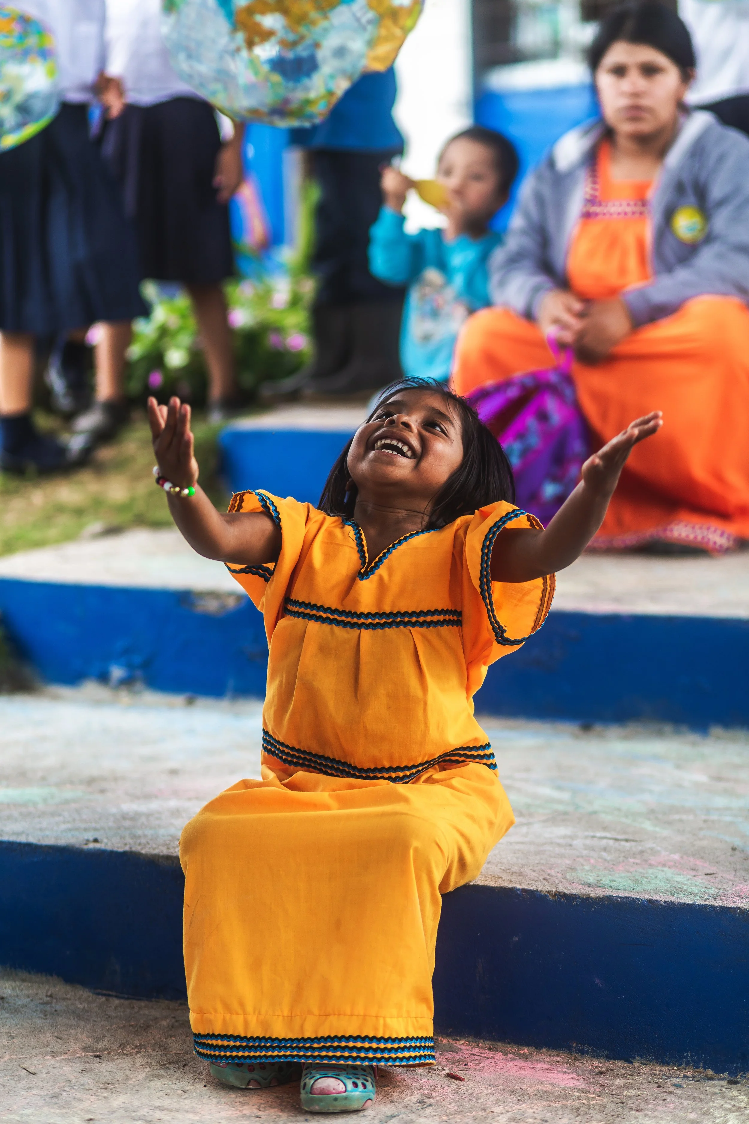 Young girl sitting on the ground, smiling and reaching up with her arms. She is dressed in a bright orange traditional dress with black, orange, and blue embroidery. Behind her, a woman and a young boy are sitting and standing, respectively, with the boy drinking from a banana. The background shows other people and colorful balloons.