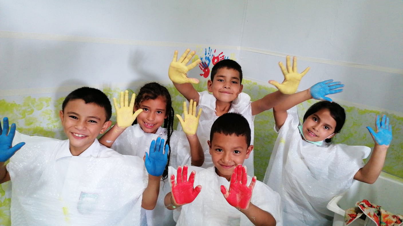 Six children smiling and showing their hands painted in blue, red, and yellow, celebrating in a room with painted walls.