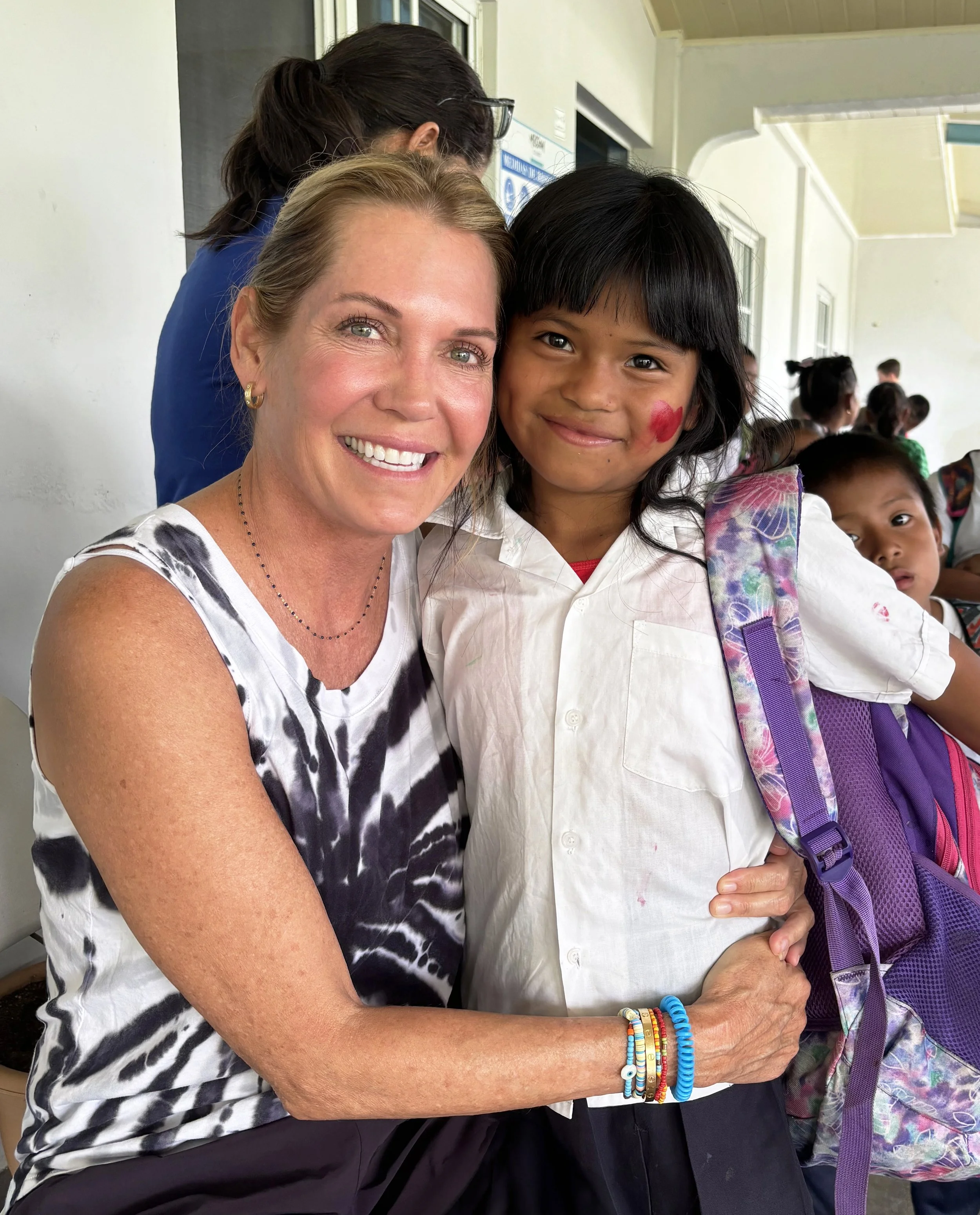 A woman and a young girl are smiling and hugging each other. The woman has light skin and light-colored hair and is wearing a sleeveless black and white patterned top, with colorful bracelets on her wrist. The girl, with darker skin and black hair, has face paint of a red heart on her cheek, and is wearing a white shirt and a purple backpack. They are indoors, with several other children and an adult visible in the background.
