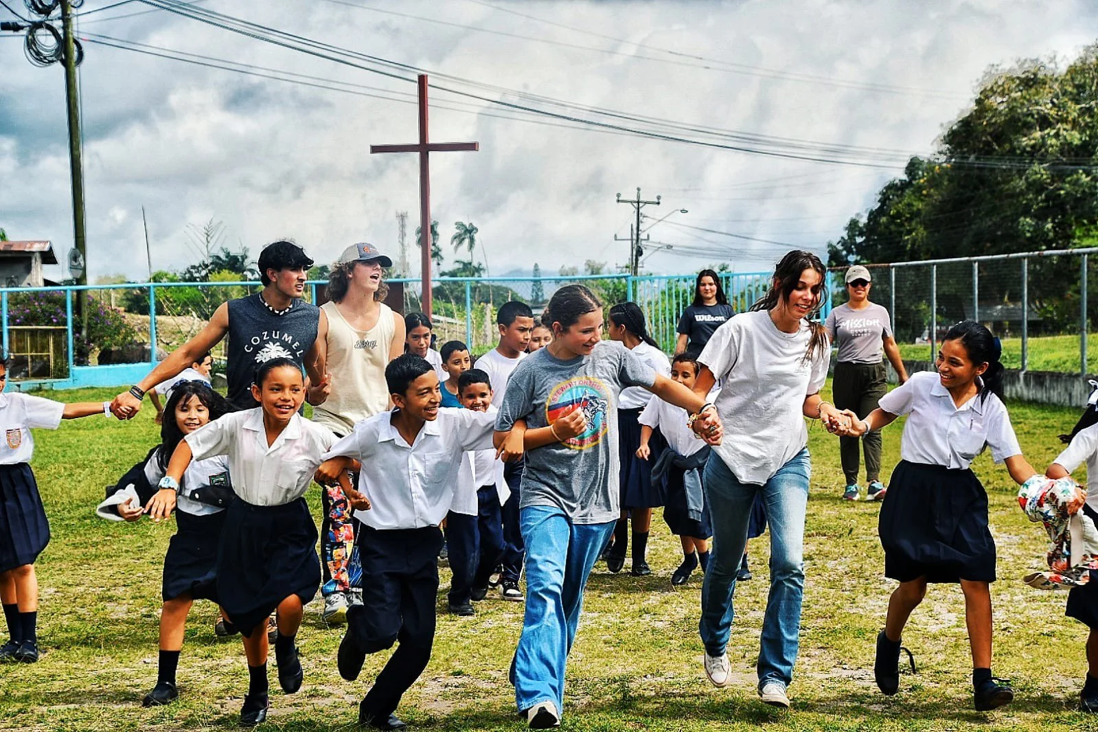 Group of children and adults enjoying outdoor activity on a grassy field, holding hands and running together, with a fence and trees in the background.