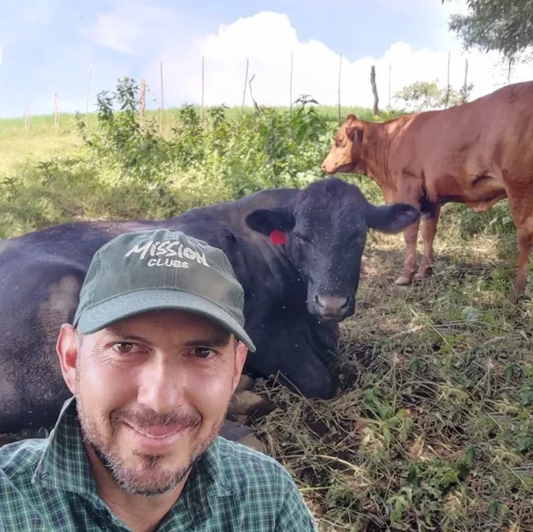 A man taking a selfie outdoors with two cows in the background, one lying down and one standing, in a grassy field with a partly cloudy sky.