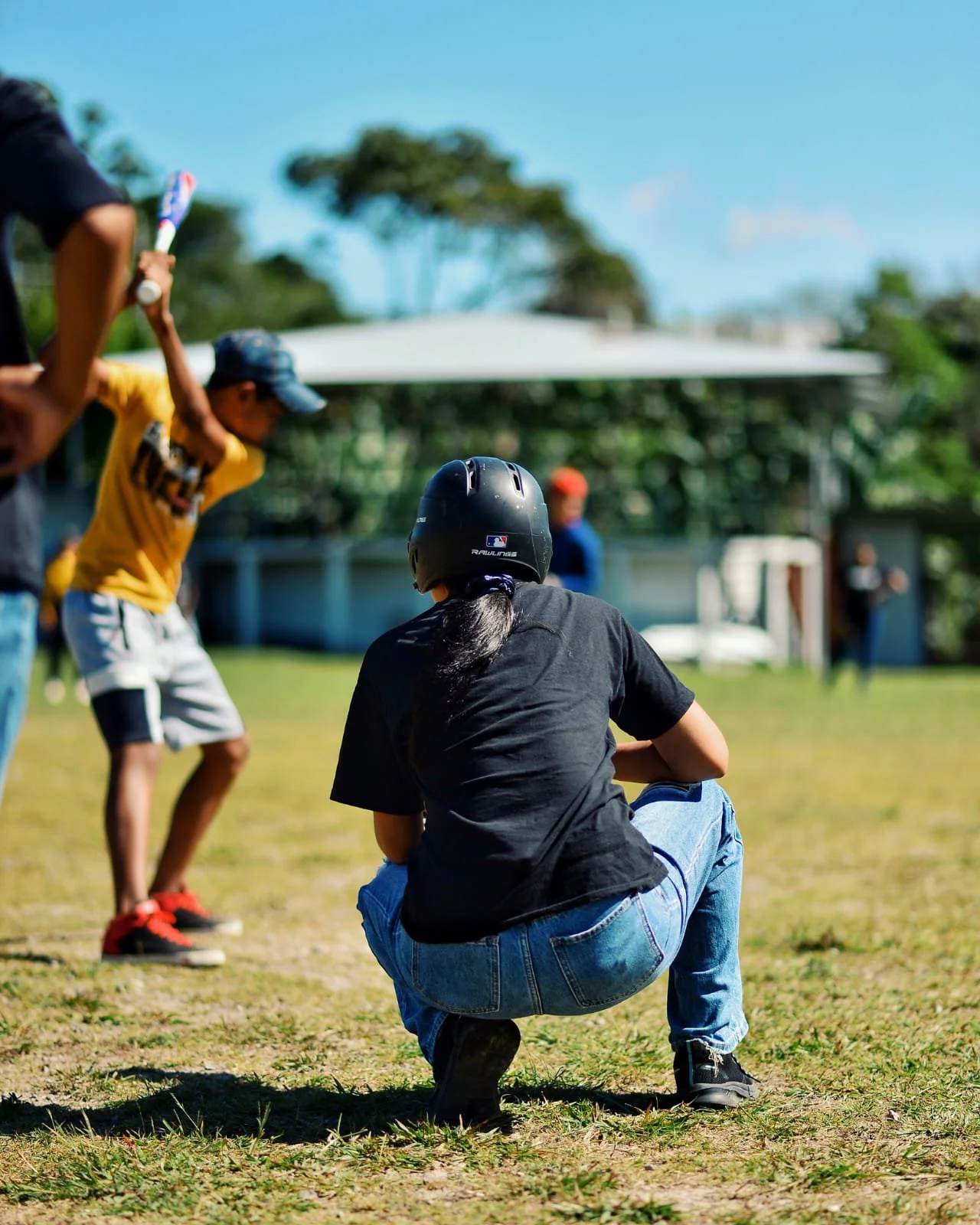 A person in a black t-shirt and blue jeans is squatting on the grass, wearing a black helmet. In the background, children are playing baseball or softball, with one child in a yellow shirt holding a bat, another child appears to be running, and a person in a blue shirt and red hat is also visible. There are trees and a building with a canopy in the distance under a clear blue sky.