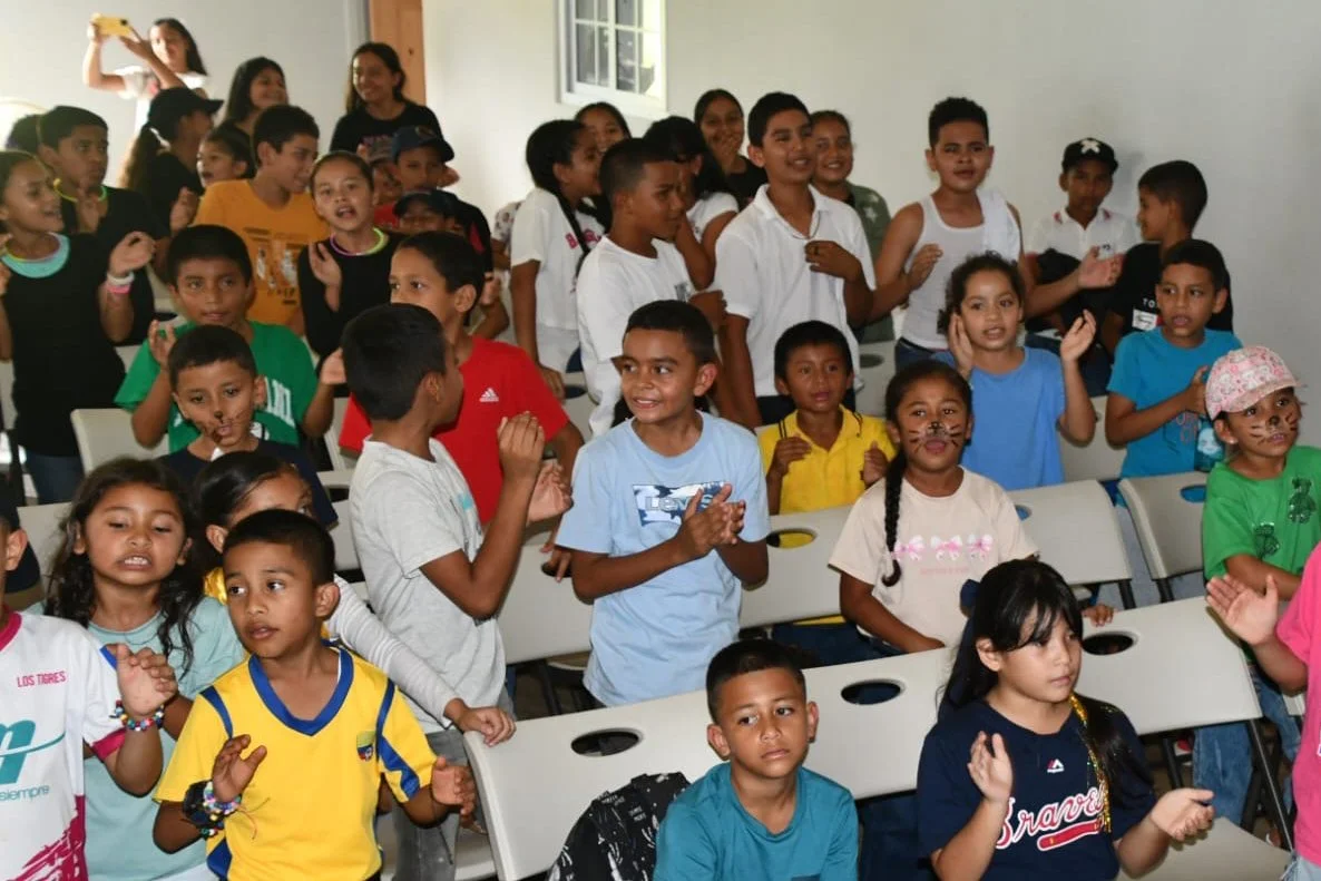 Group of children and teenagers gathered in a classroom or community center, some are standing and clapping or singing, with a few adults in the background.