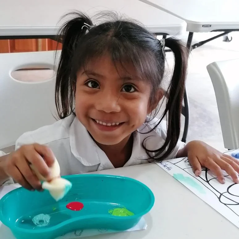 Young girl with dark hair in pigtails, smiling and playing with paint on a table.