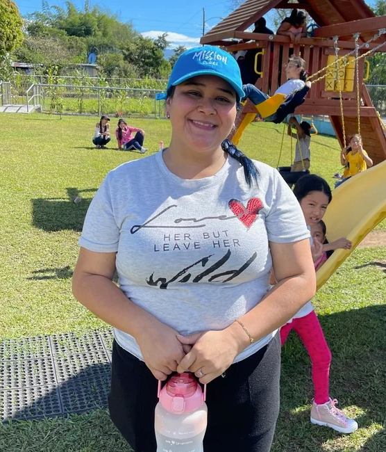 Smiling woman in a gray t-shirt and blue cap standing in a playground with children playing on a slide and swing set in the background.