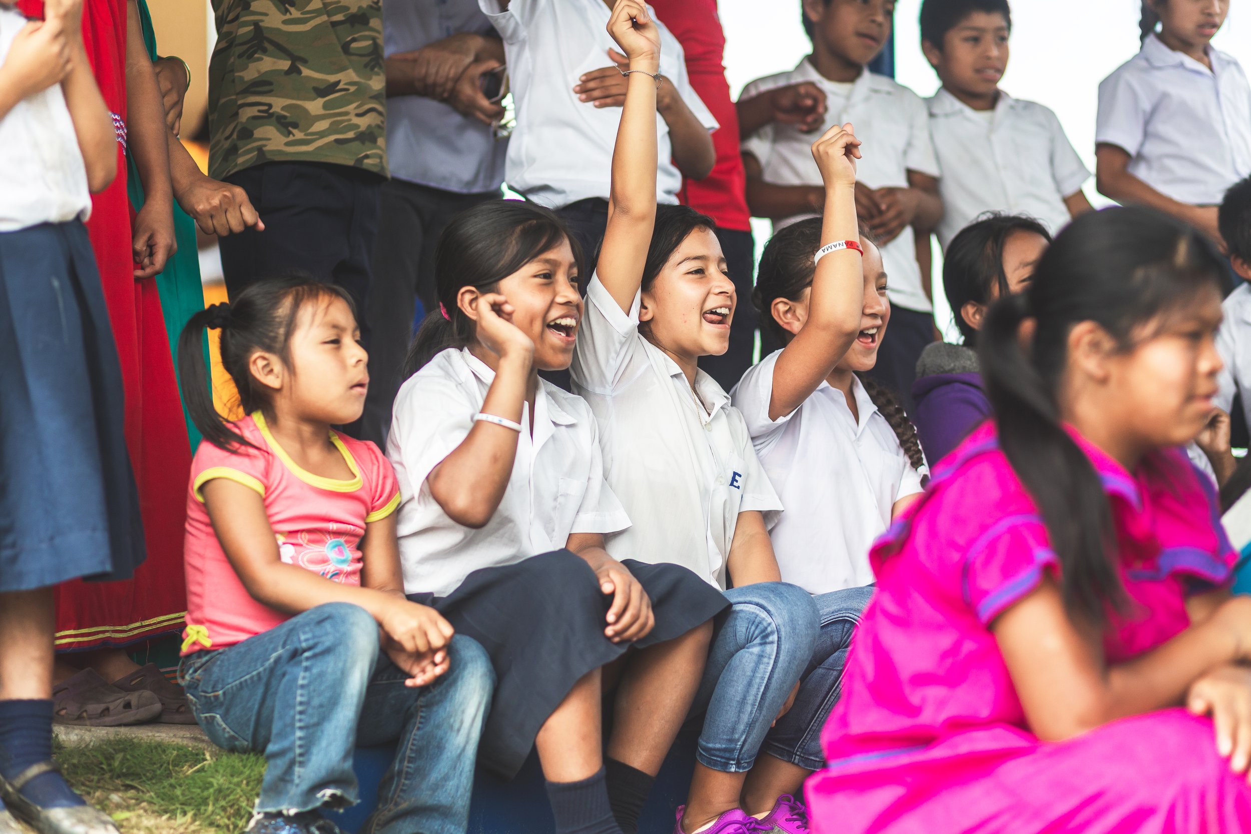 Group of children sitting outdoors, with some raising hands, engaged in an activity or discussion, smiling and appearing happy.