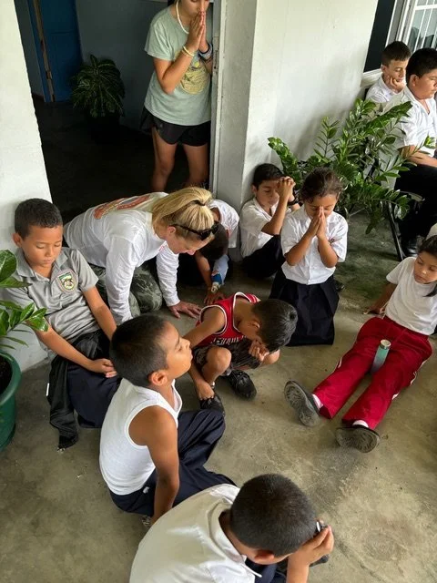 Children sitting on the floor and praying with a teacher standing behind them, outdoors near a building and green plants.