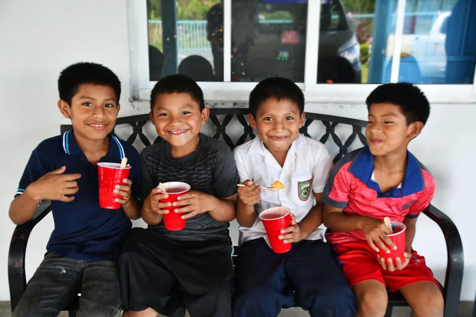 Four young boys sitting on a bench, smiling, and holding red cups with a drink, enjoying each other's company.