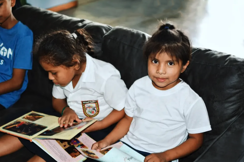 Two young girls sitting on a black sofa, reading magazines. One girl looks at the camera and the other is focused on her magazine. Both girls are wearing white shirts.