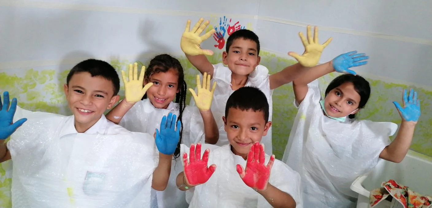 Six children with colorful painted hands smiling and waving in a classroom, wearing white smocks.