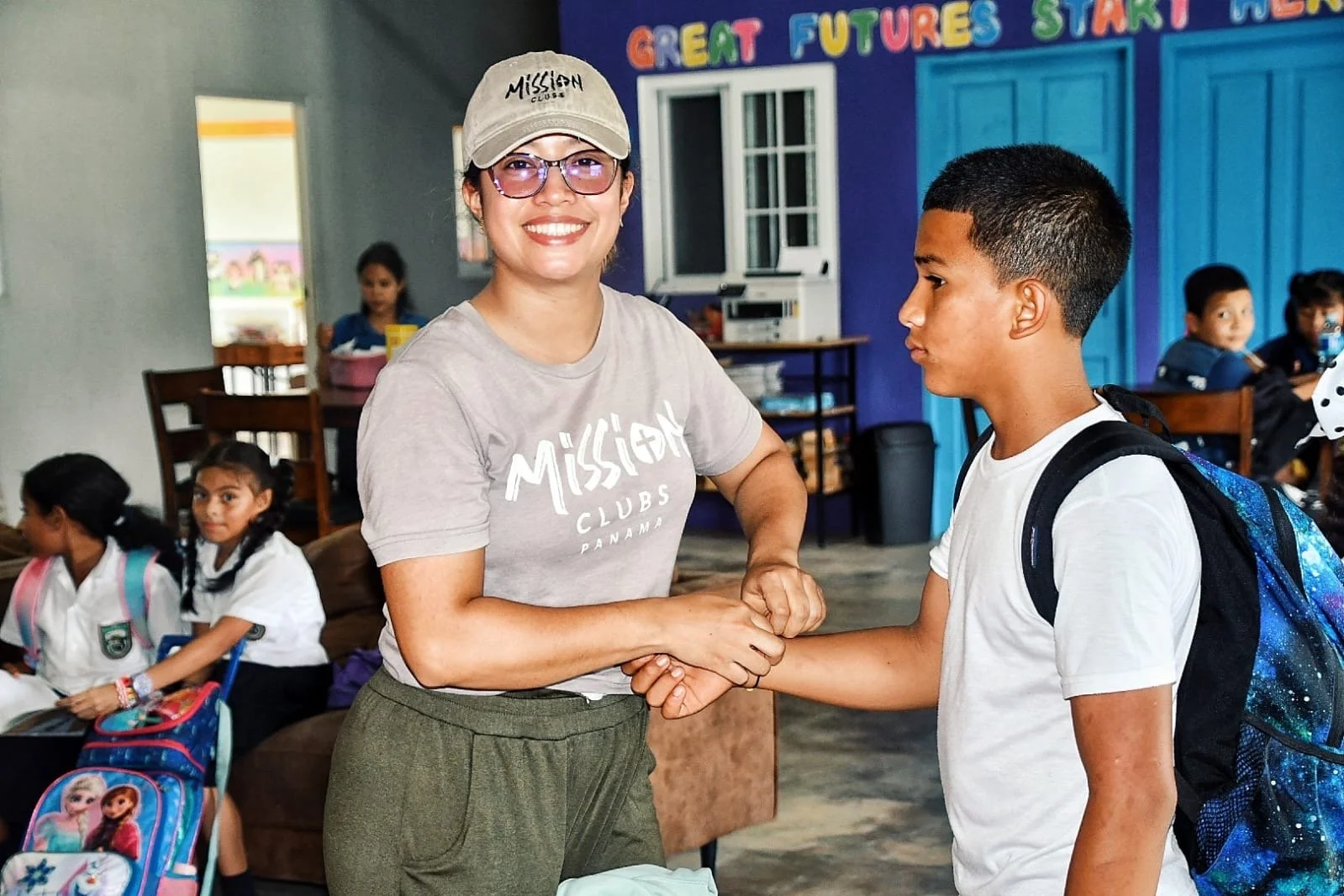 A woman smiling and shaking hands with a young boy with a backpack inside a classroom. Several other children are sitting with backpacks, and a colorful wall with the words 'Great Futures Start Here' is visible in the background.