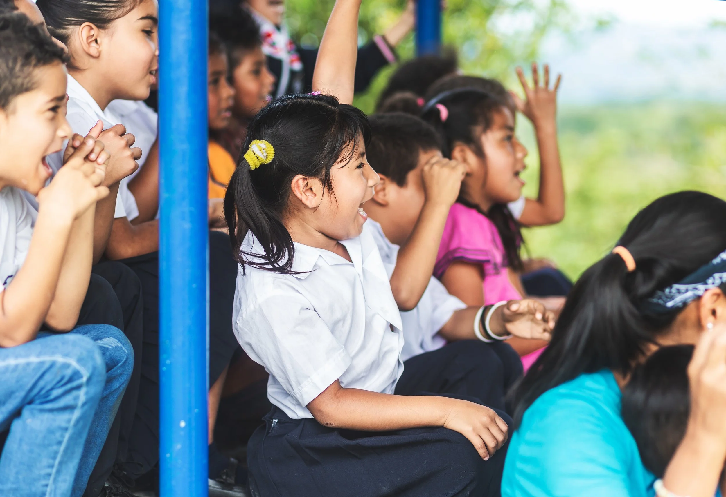 A group of diverse children sitting outdoors with trees in the background, some raising hands, engaged and smiling.