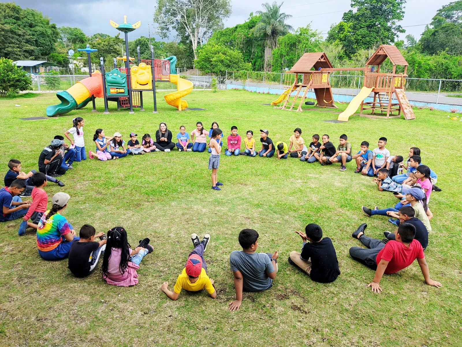 Group of children and adults sitting in a large circle on a grassy field at a playground, with playground equipment including slides and climbing structures in the background.