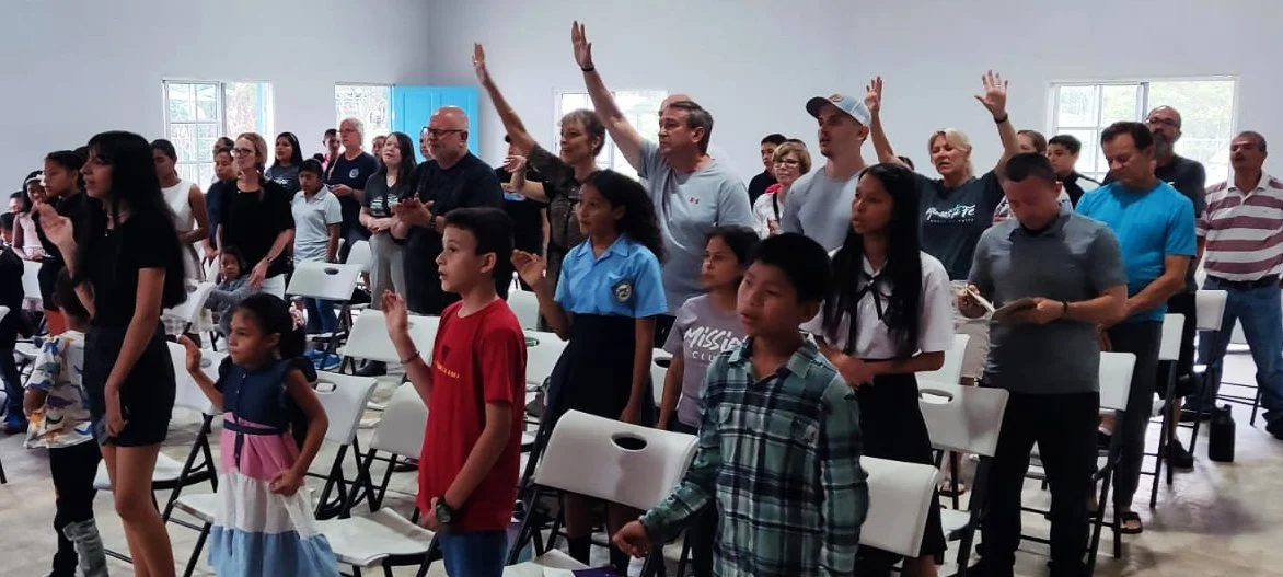 Group of diverse people standing and raising hands in a bright, spacious room with large windows, some sitting on chairs, engaged in a gathering or event.
