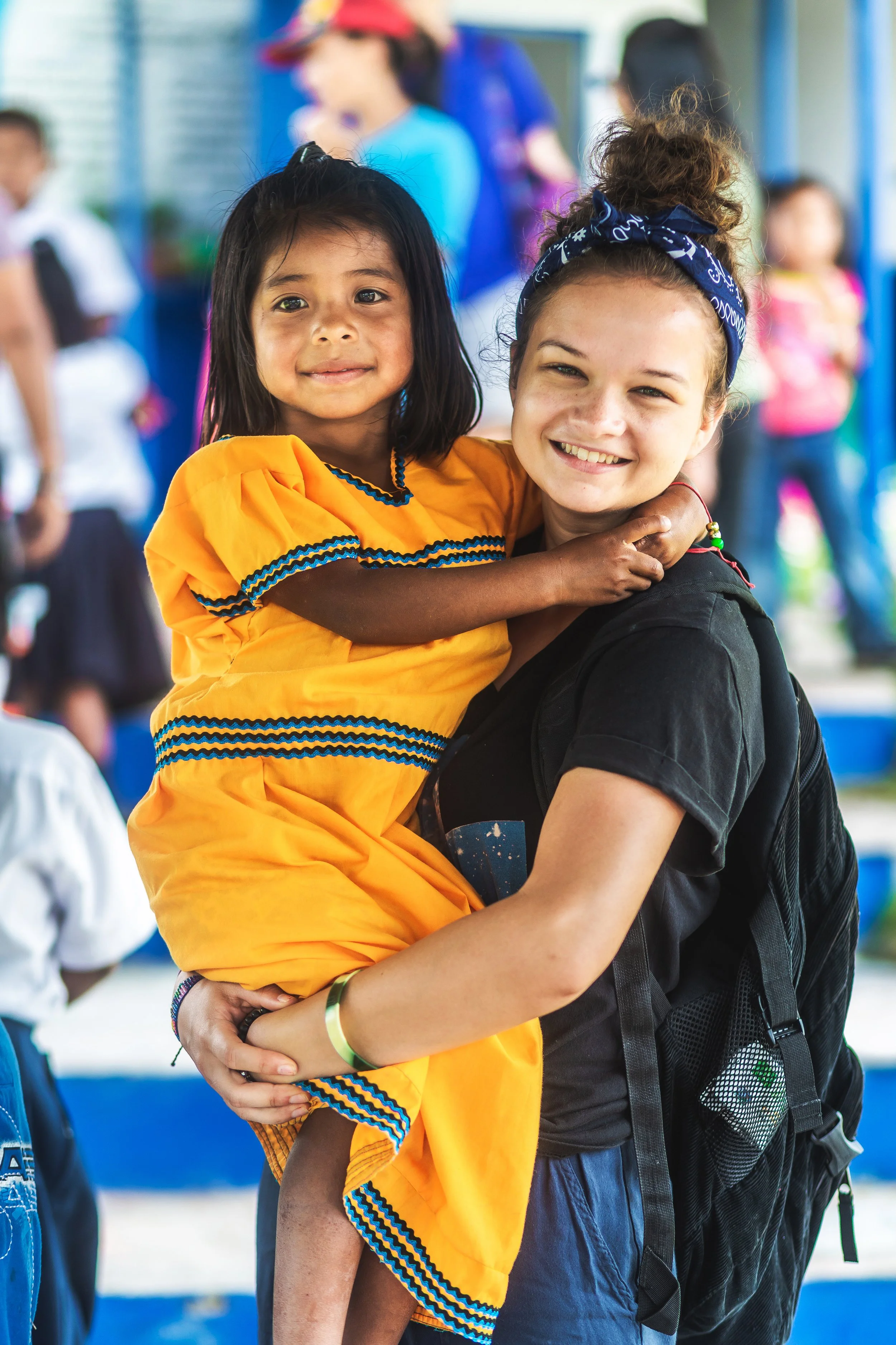 A young girl in a yellow dress being held by an older girl or woman with a backpack, smiling at the camera. The background shows other children and adults in a brightly colored environment, possibly a school or community center.