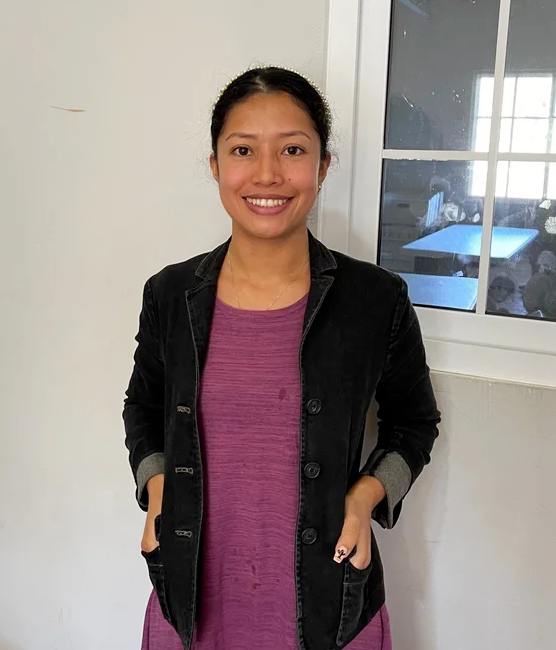A young woman with dark hair tied back, smiling, wearing a black jacket over a purple dress, standing near a window in a room with white walls.