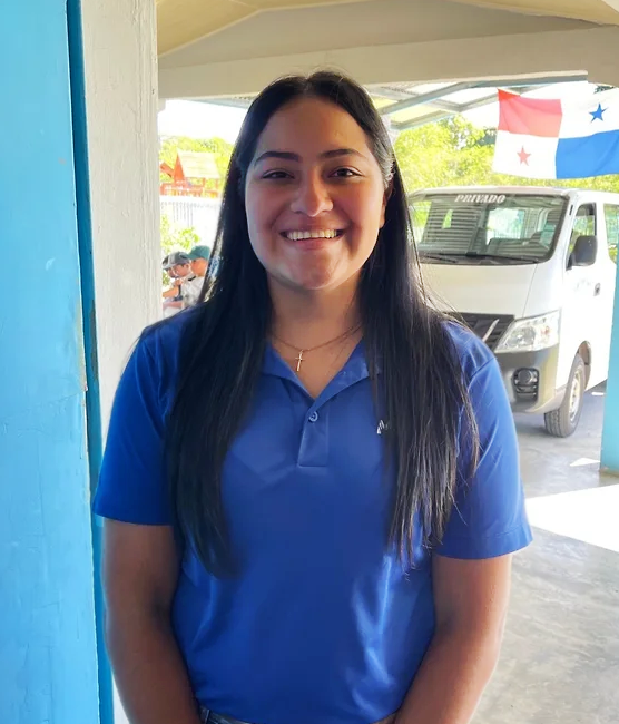 Smiling woman with long dark hair wearing a blue polo shirt standing outdoors with a Texas flag and a white van in the background.