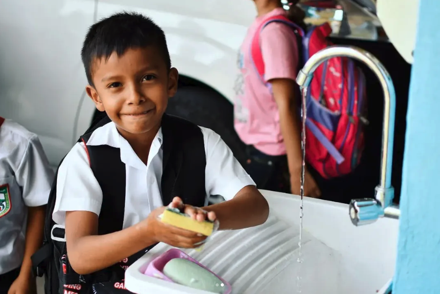 A young boy in a school uniform washing his hands at a sink outside, with children and backpacks in the background.