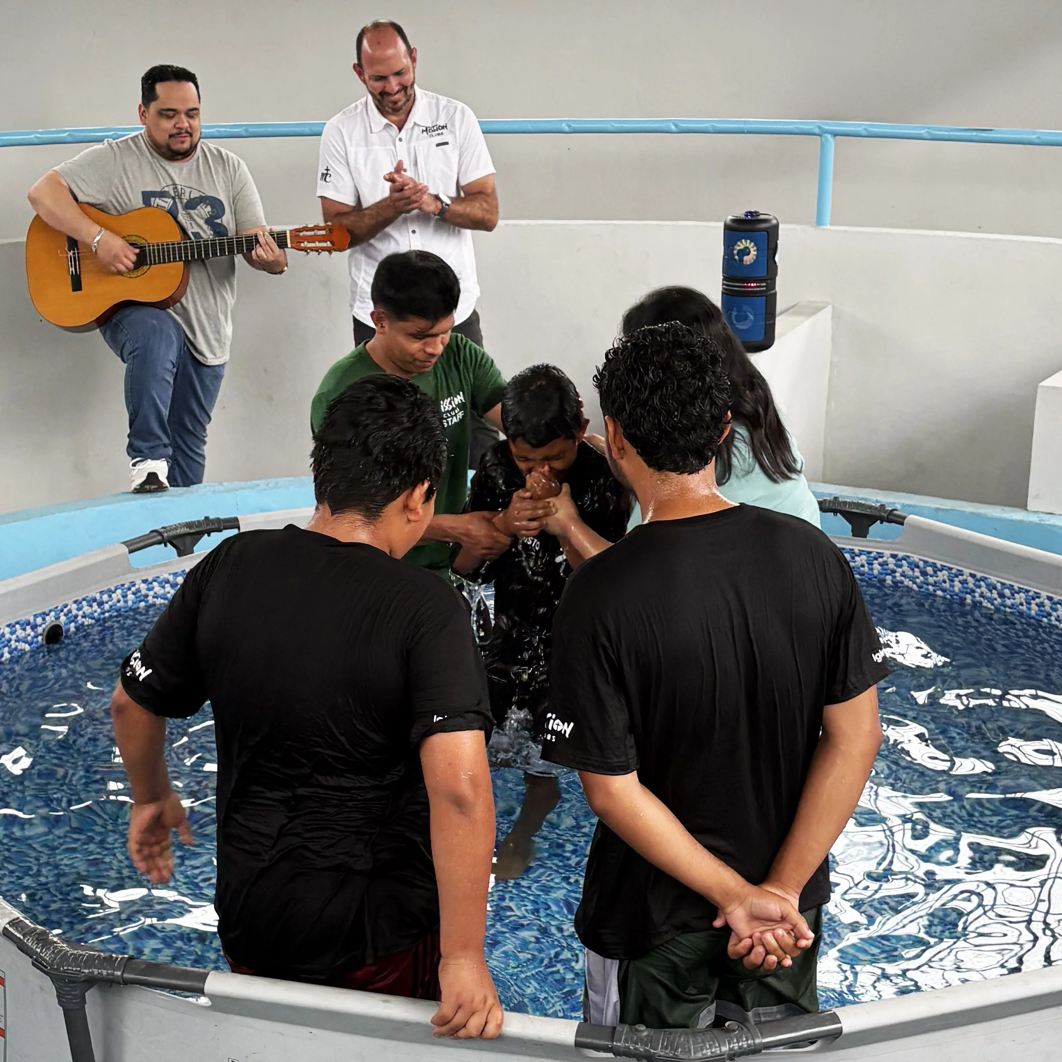 People participating in a religious baptism ceremony in a small pool, with a guitarist and a pastor present.