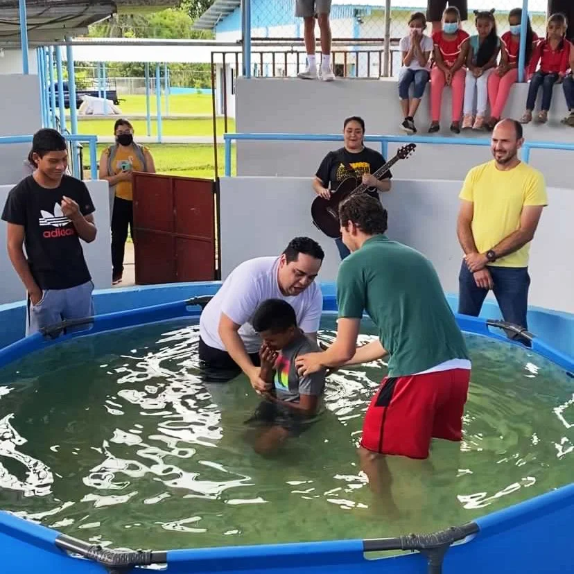 Children and adults participating in a baptism ceremony in a small pool, with a person being baptized, overseen by others, in an outdoor setting with onlookers and a guitarist in the background.