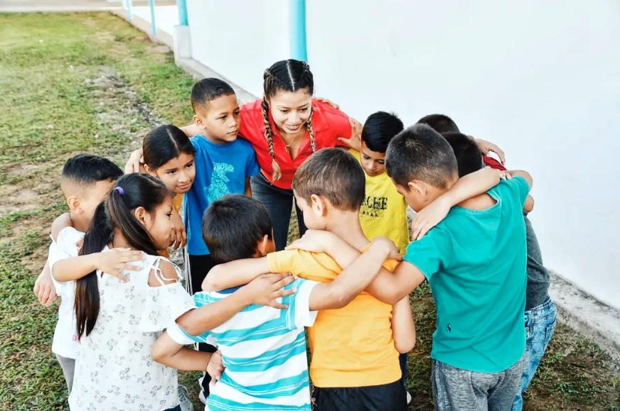 A group of children and a woman in a huddle outdoors, hugging and smiling.