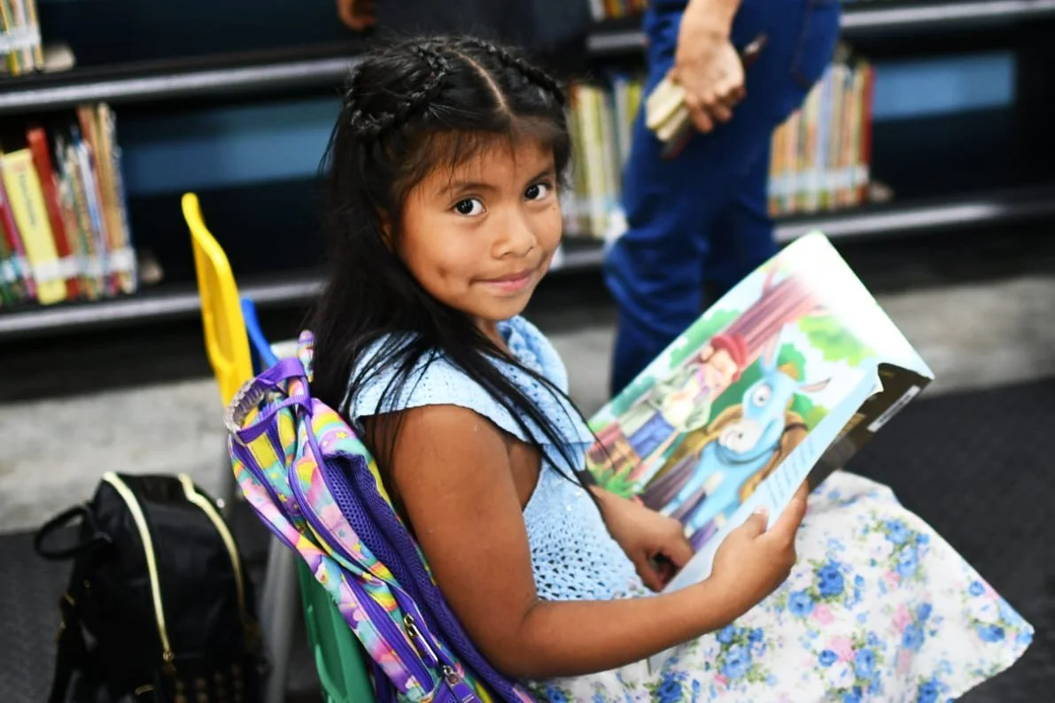 A young girl sitting in a library, holding a colorful children's book featuring animals, with a purple backpack on her back and a yellow chair nearby.