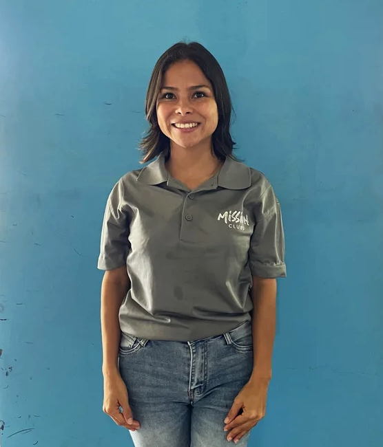 Woman in gray polo shirt and jeans standing against a blue wall, smiling.
