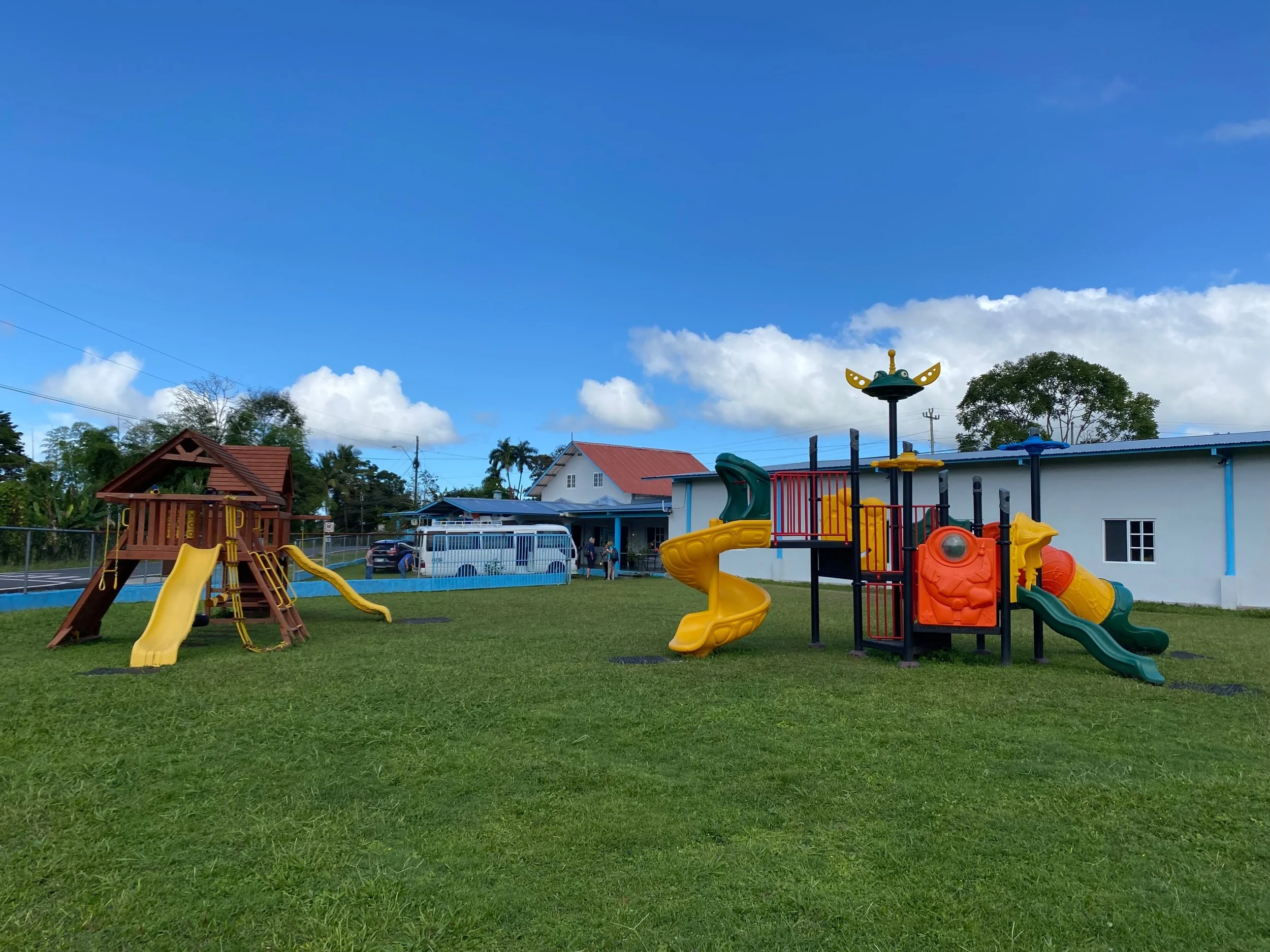 A playground with various slides, a climbing structure, and a merry-go-round, situated on a grassy field with houses, trees, and a blue sky with clouds in the background.