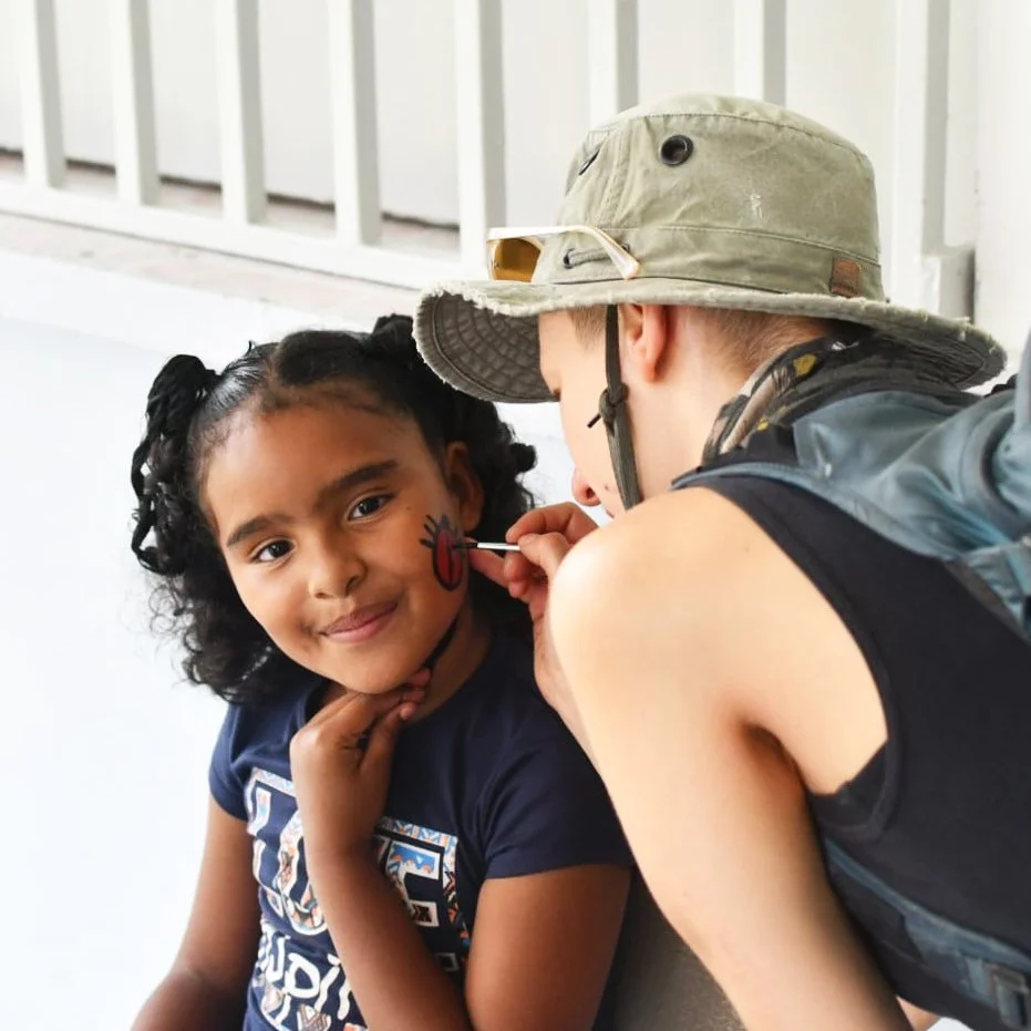 A young girl with dark curly hair has her face painted with a red ladybug and is smiling while a face painter paints the ladybug on her cheek. The face painter is wearing a wide-brimmed hat and a backpack.