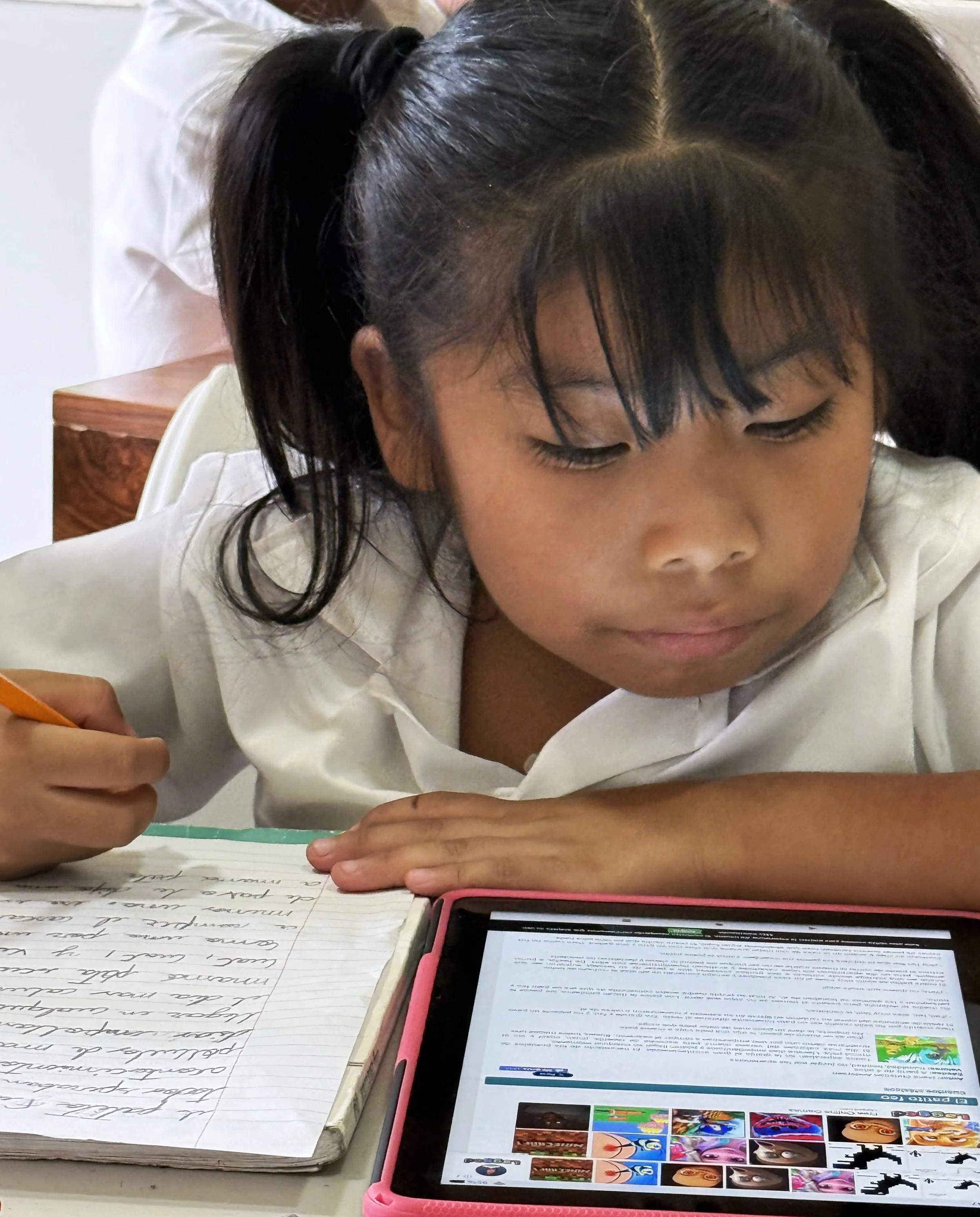 A young girl with black hair in pigtails is sitting at a table, writing in a notebook. Her face is partially visible, and she appears to be focused on her work. There is a tablet with a colorful display and text on the screen in front of her, along with a notebook filled with handwritten notes.