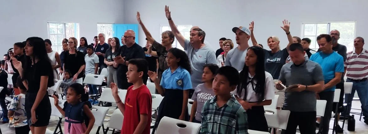 People standing and raising their hands in a spacious room with white walls and multiple windows.