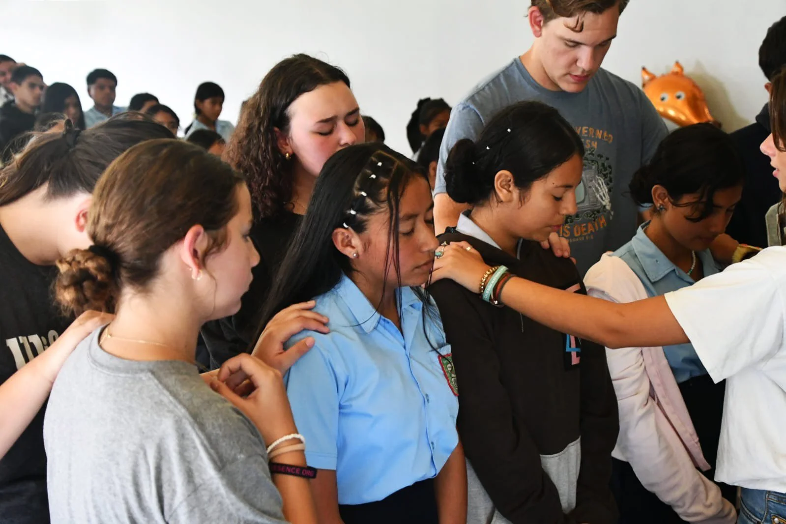 A diverse group of students in school uniforms praying with heads bowed and hands placed on shoulders in a classroom.