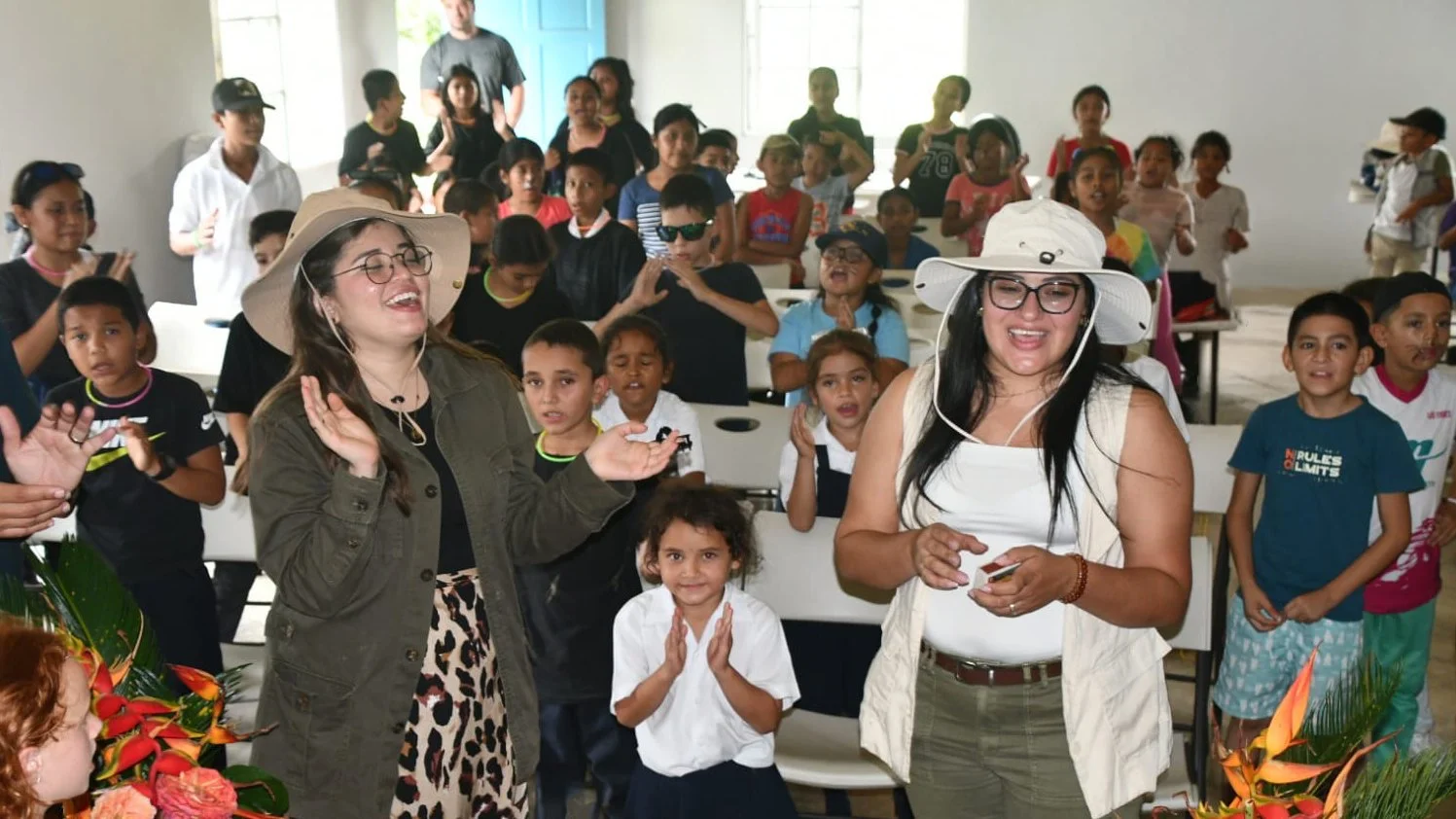 A group of children and two women wearing sun hats in a bright room, some children are singing or clapping, with a joyful atmosphere.