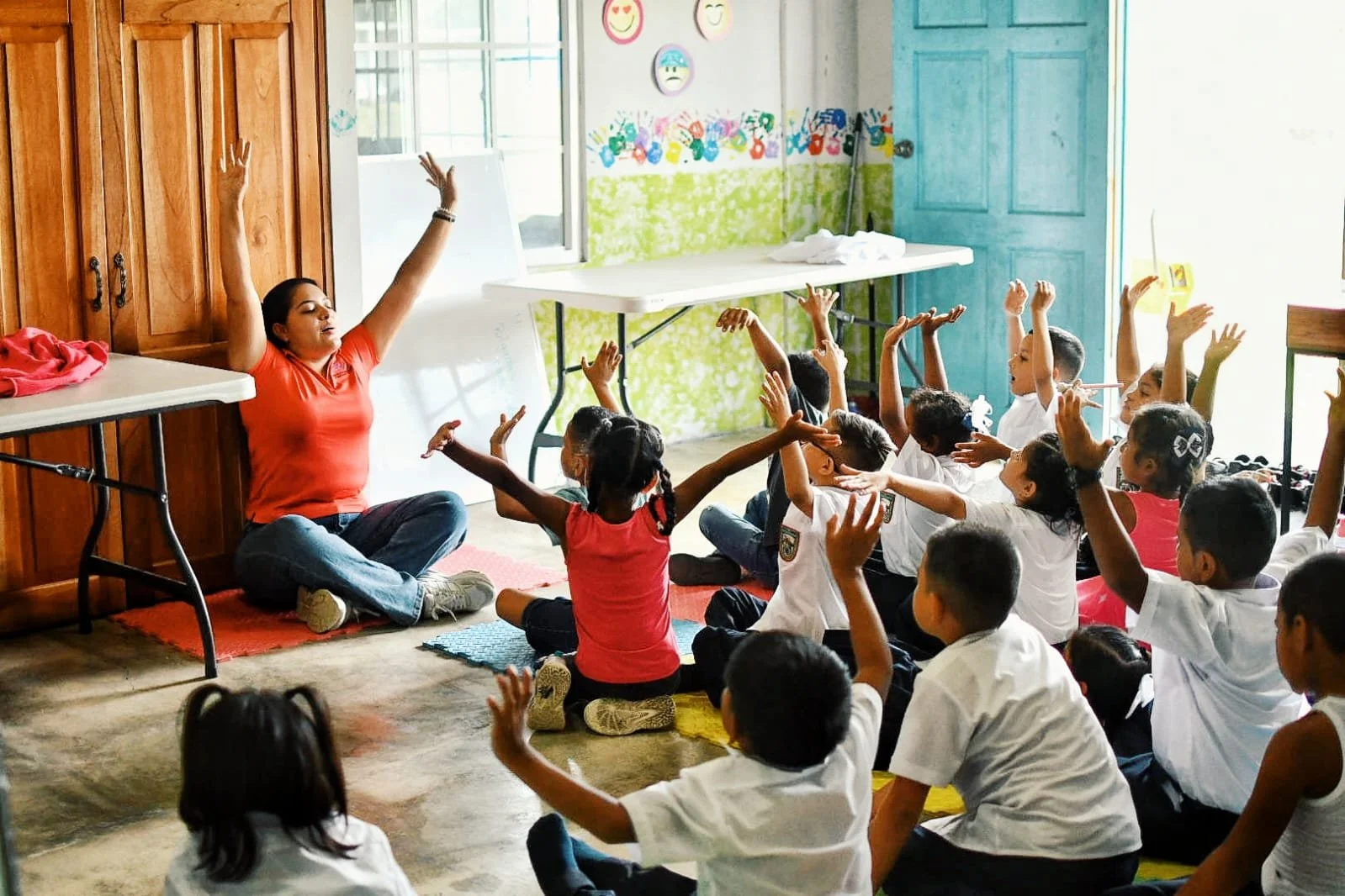 A woman leading a seated yoga or meditation session with children raising their hands in a classroom or community center, colorful wall decorations, and bright natural light.