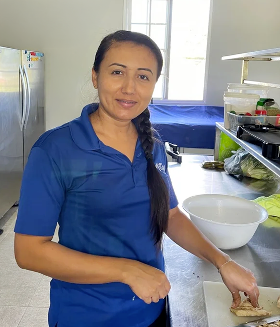 A woman in a blue polo shirt standing in a kitchen, slicing a piece of meat on a cutting board, with a white mixing bowl and various kitchen supplies nearby.