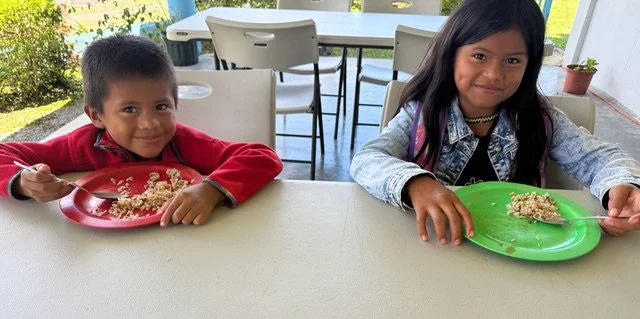 Two children sitting at a table eating rice from colored plates, one boy with a red shirt and a girl with a denim jacket, outdoors with patio furniture and greenery in the background.