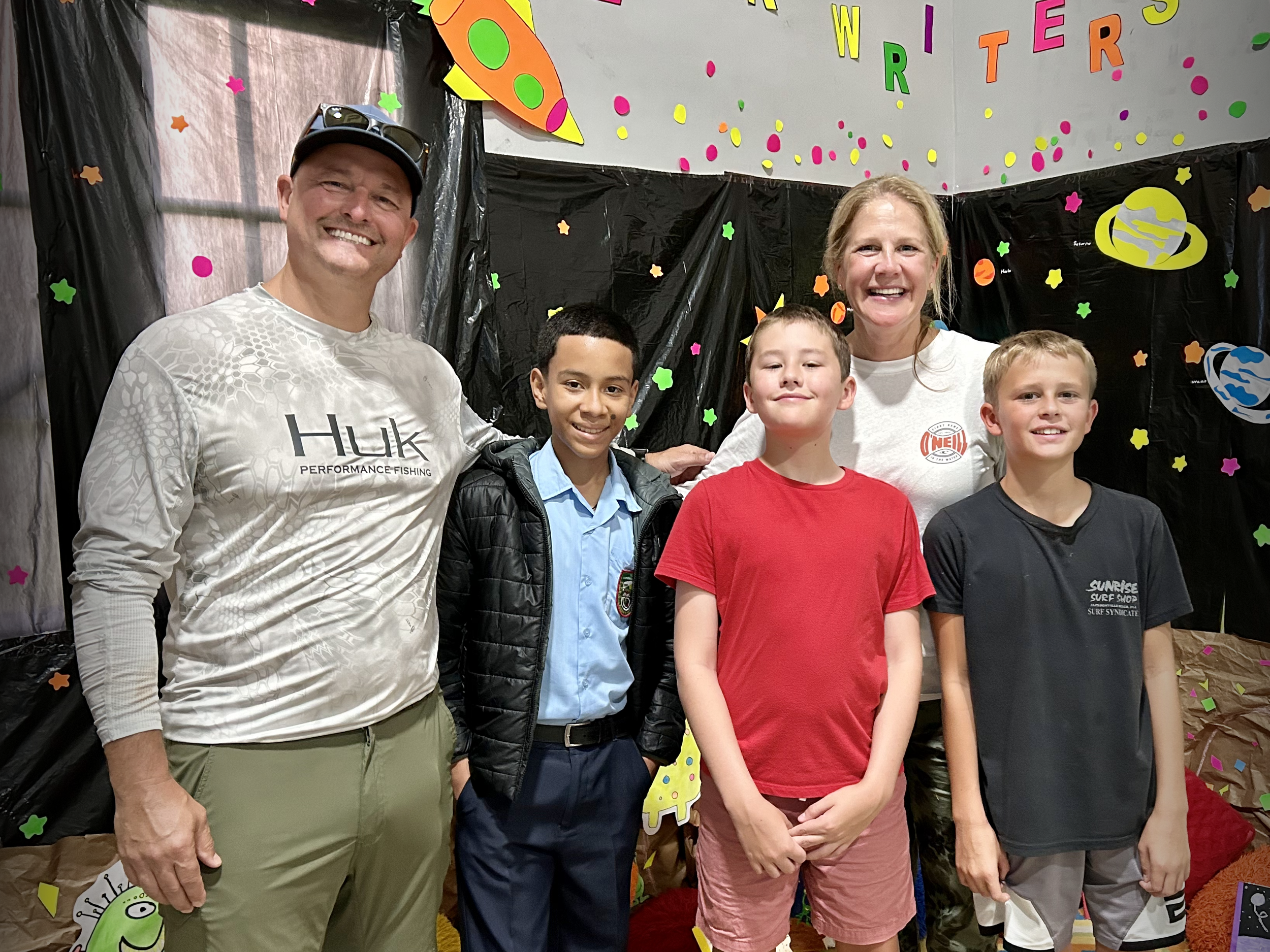 Group of five smiling people standing in front of space-themed decorations with colorful paper stars, planets, and a rocket ship, celebrating a party or event.