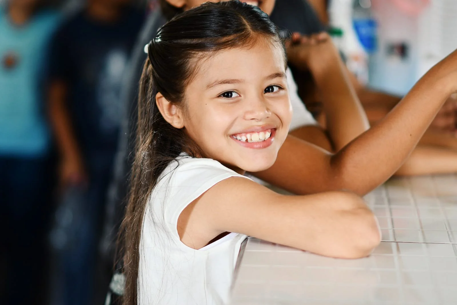 Smiling young girl with long brown hair sitting at a tiled counter in a classroom or cafeteria.
