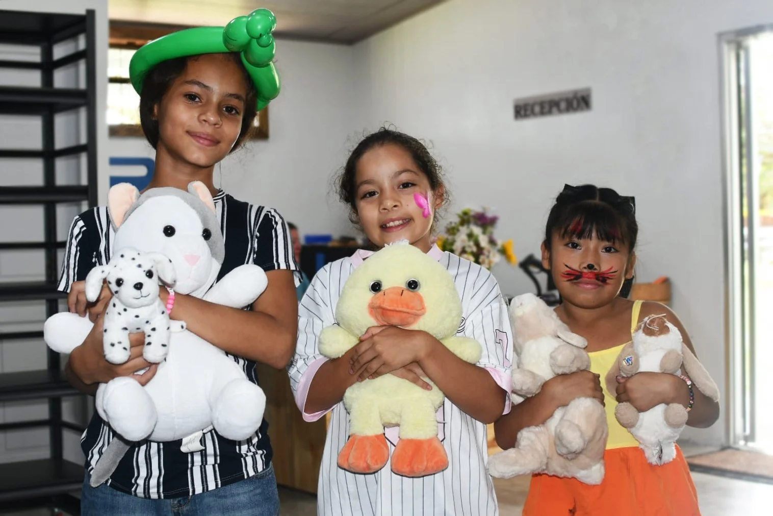 Three young girls standing indoors, each holding stuffed animals. The girl on the left is wearing a balloon hat and holding a large white stuffed animal and a smaller leopard plush. The girl in the middle has a painted pink flower on her face and holds a yellow duck plush. The girl on the right has face paint resembling a cat's nose and whiskers and holds a tan teddy bear plush. They are smiling and appear to be at a playful event or party.