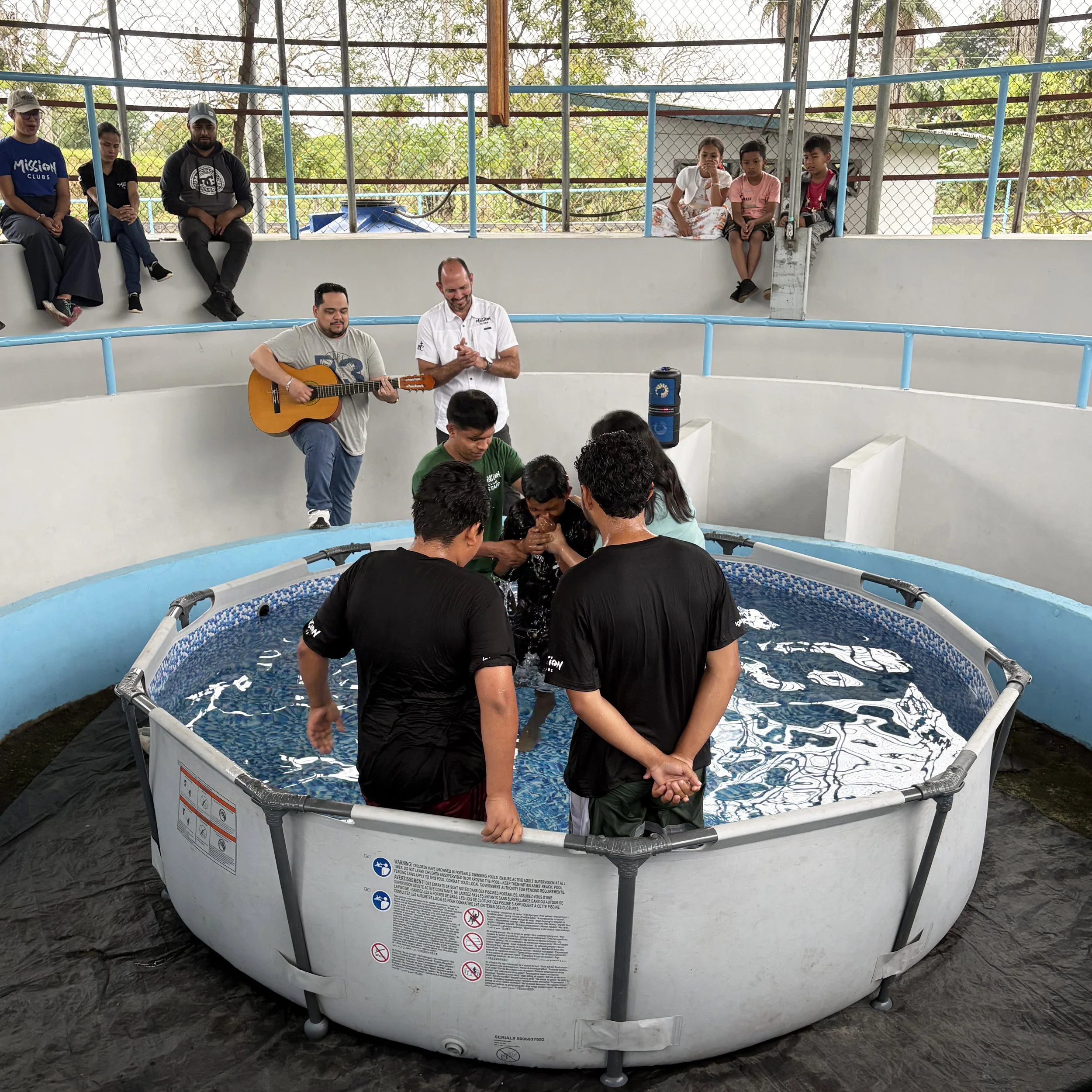 A group of children and adults standing and sitting around a circular baptism pool during a baptism ceremony outdoors, with a chain-link fence and trees in the background.