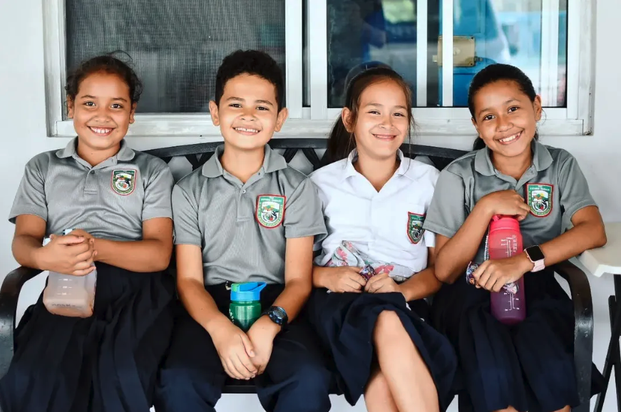 Four young students sitting on a bench, smiling, wearing school uniforms, holding water bottles, in a classroom setting.