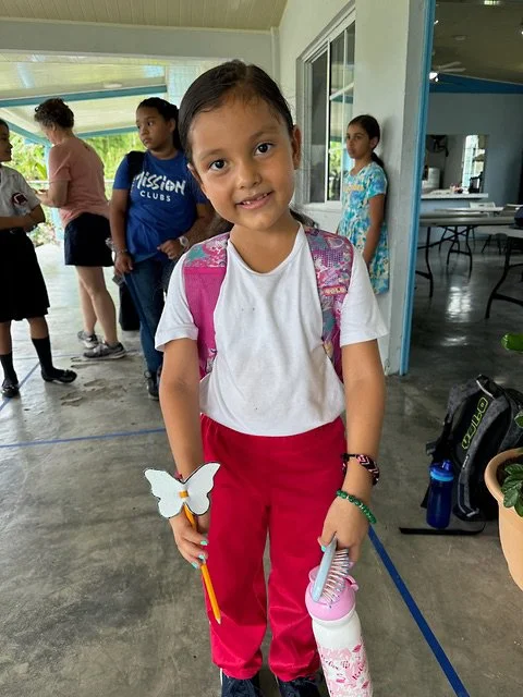 A young girl with long dark hair, wearing a white t-shirt and red pants, standing indoors with a pink backpack. She is holding a butterfly-shaped flute and a water bottle, smiling at the camera. In the background, several other children are standing near a window and doorway.
