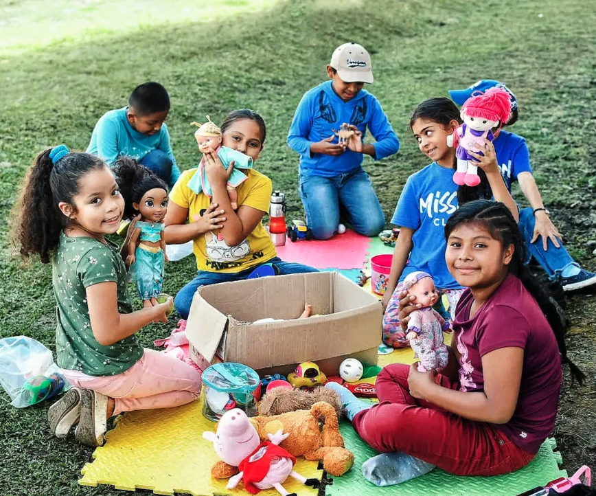 Group of children playing with dolls and toys outdoors on a grassy area.