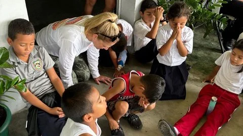 A group of young children and a woman, possibly a teacher, are kneeling or sitting on the floor in a circle, appear to be praying or meditating, with some children having their hands clasped and eyes closed.