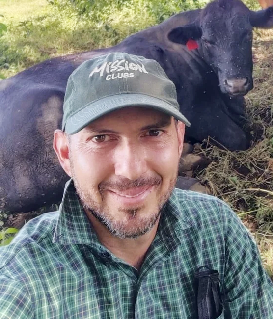 Man taking a selfie outdoors with a black calf laying behind him, all on grassy ground.