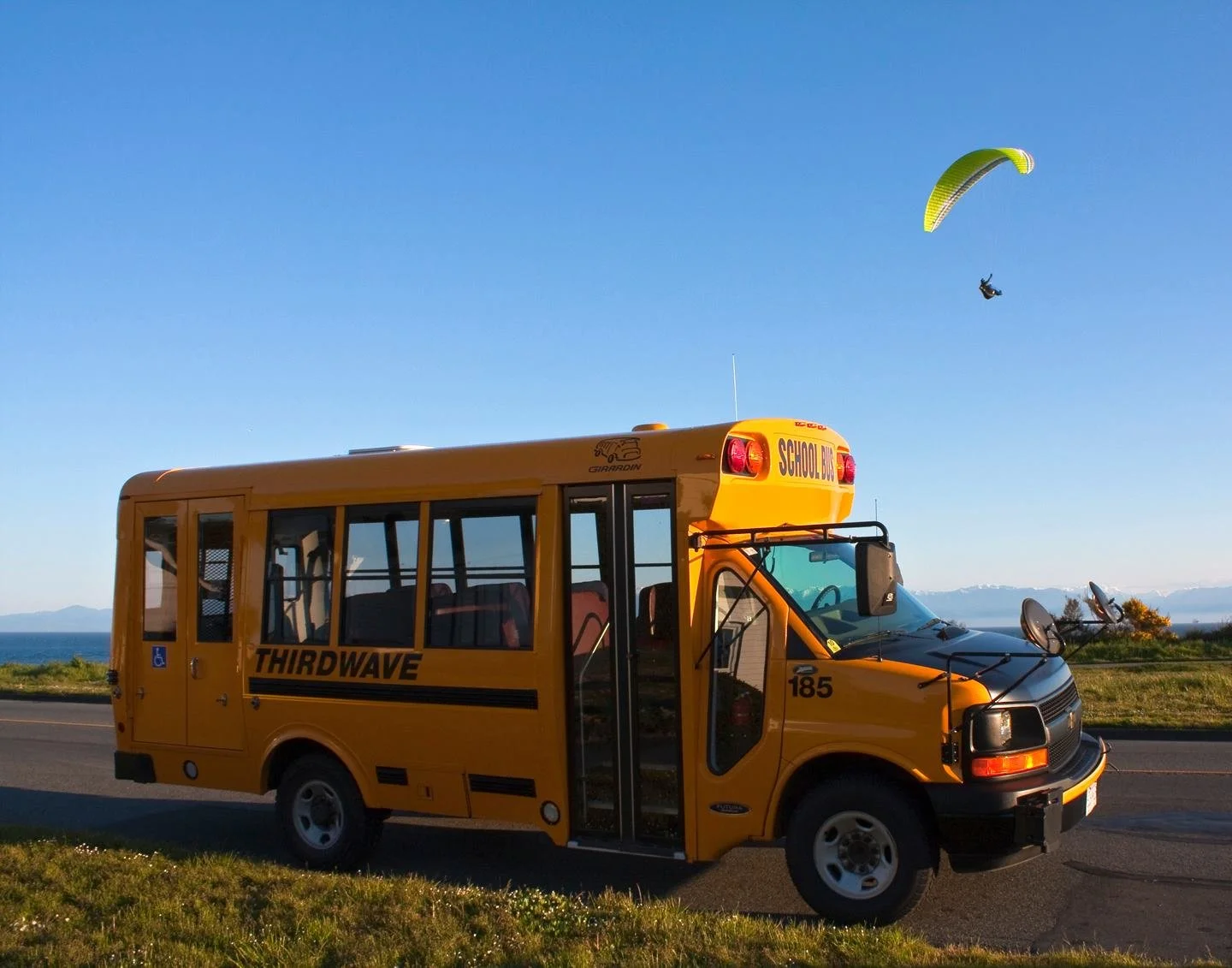 Yellow school bus labeled "ThirdWave" parked beside a road with a landscape backdrop, while a paraglider soars in a clear blue sky nearby.
