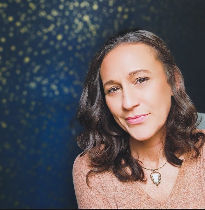 A woman with shoulder-length wavy dark hair, wearing a peach sweater and a necklace with a white enamel design, smiling softly with a slightly tilted head, against a dark background with gold specks.