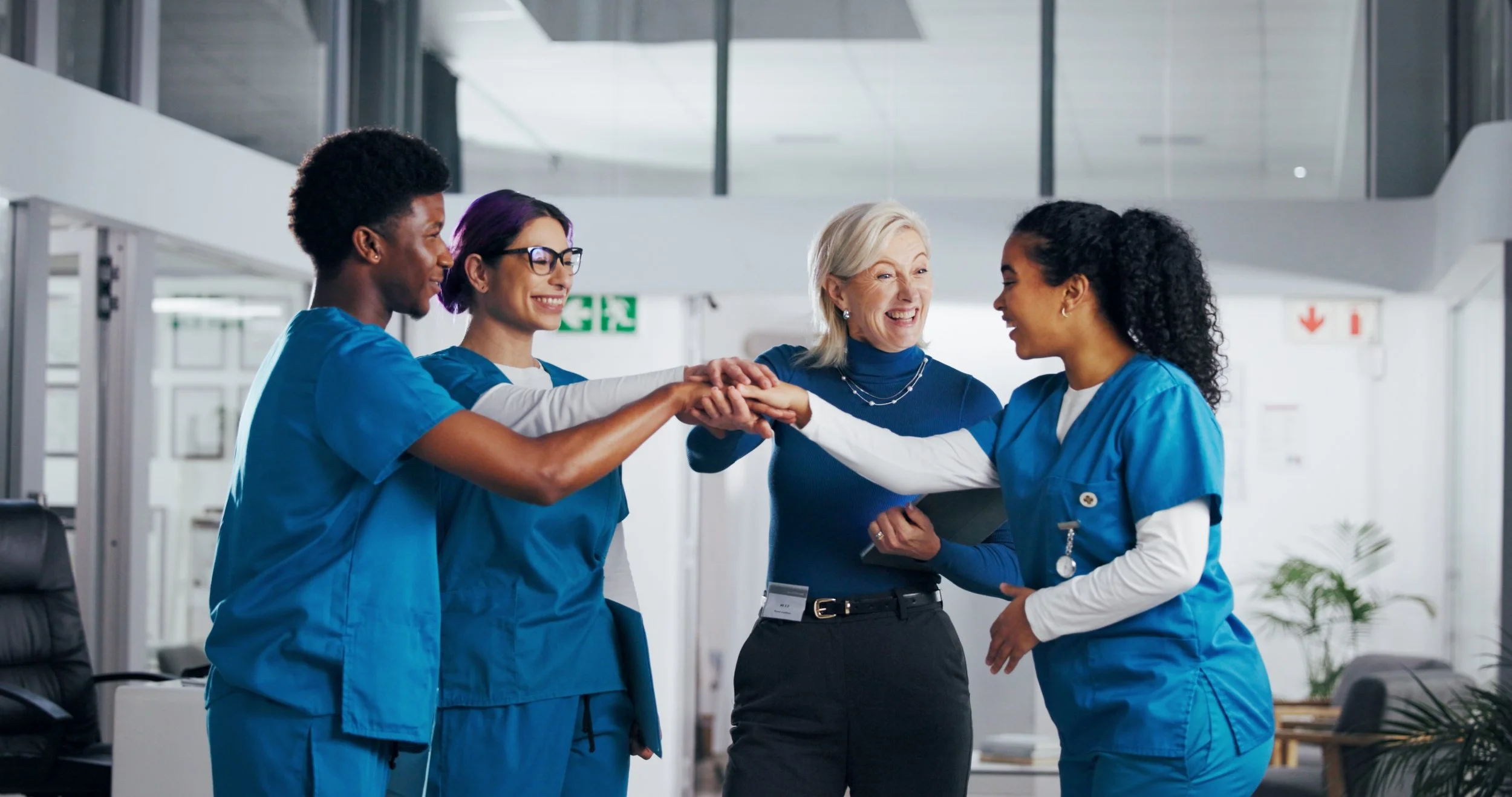 Four healthcare workers, three women and one man, in nurse scrubs reaching their hands in a circle in a hospital lobby for a team cheer or gesture of unity.