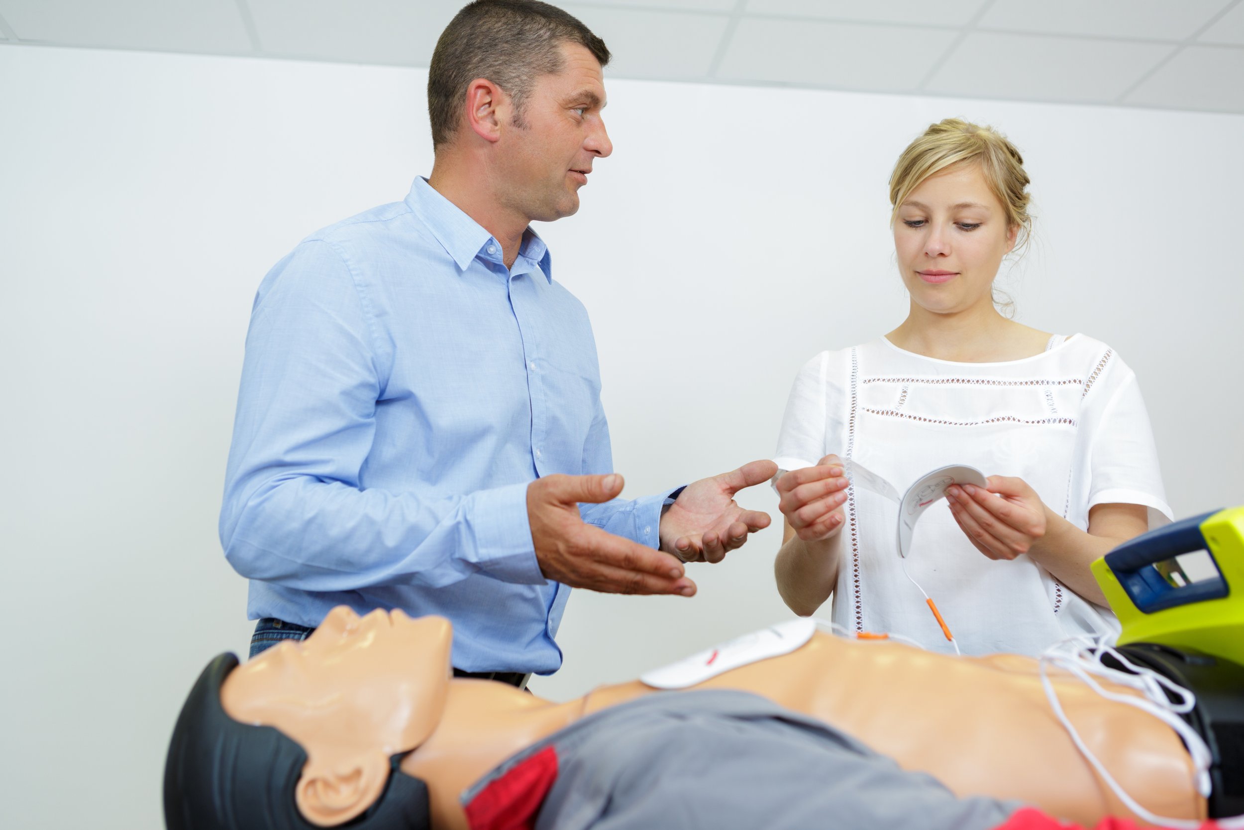 Two people, a man and a woman, are in a medical or training setting. The man, wearing a light blue shirt, is explaining something to the woman, who is holding a medical device or model. A medical mannequin or dummy is on the table in front of them, suggesting a training or demonstration scenario.